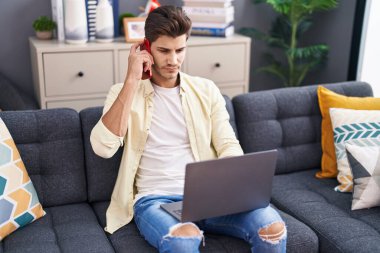 Young hispanic man talking on smartphone using laptop at home
