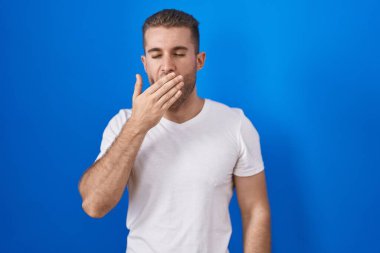 Young caucasian man standing over blue background bored yawning tired covering mouth with hand. restless and sleepiness. 