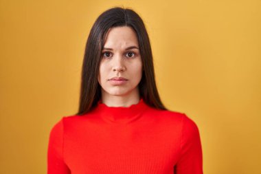 Young hispanic woman standing over yellow background relaxed with serious expression on face. simple and natural looking at the camera. 