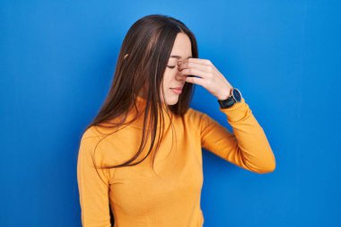 Mavi arka planda duran esmer kadın yorgun burnu ve gözleri ovuşturup yorgun ve baş ağrısı çekiyor. Stres ve hüsran kavramı. 