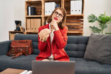 Young brunette woman at consultation office holding brain serious face thinking about question with hand on chin, thoughtful about confusing idea 