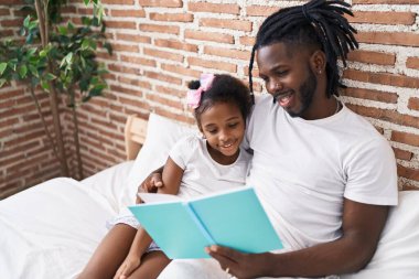 Father and daughter reading book sitting on bed at bedroom