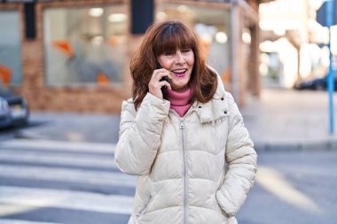 Orta yaşlı bir kadın gülümsüyor. Sokakta akıllı telefondan konuşuyor.