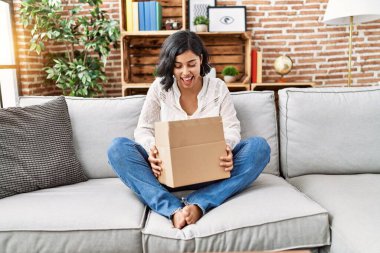 Young hispanic woman sitting on the sofa checking delivery package smiling and laughing hard out loud because funny crazy joke. 