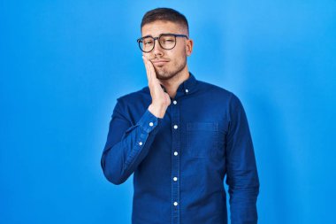 Young hispanic man wearing glasses over blue background touching mouth with hand with painful expression because of toothache or dental illness on teeth. dentist 