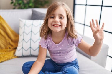 Adorable hispanic girl saying hello with hand sitting on sofa at home