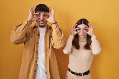 Young hispanic couple standing over yellow background trying to open eyes with fingers, sleepy and tired for morning fatigue 