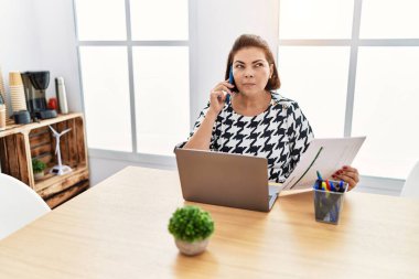 Middle age hispanic woman working at the office with laptop speaking on the phone smiling looking to the side and staring away thinking. 