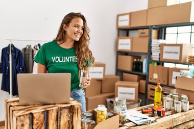 Young beautiful hispanic woman volunteer using laptop holding canned food at charity center