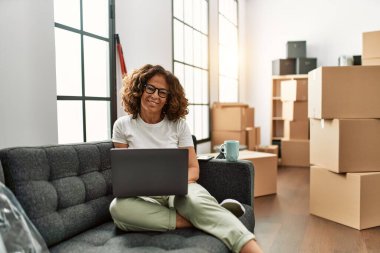 Orta yaşlı İspanyol kadın yeni evinde dizüstü bilgisayar kullanarak gülümsüyor.