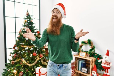 Redhead man with long beard wearing christmas hat by christmas tree smiling cheerful offering hands giving assistance and acceptance. 