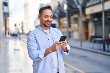 Sokakta akıllı telefon kullanan, kendinden emin gülümseyen beyaz bir genç.