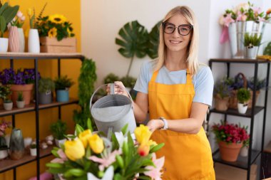 Young blonde woman florist smiling confident watering plant at flower shop