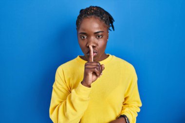 Beautiful black woman standing over blue background asking to be quiet with finger on lips. silence and secret concept. 