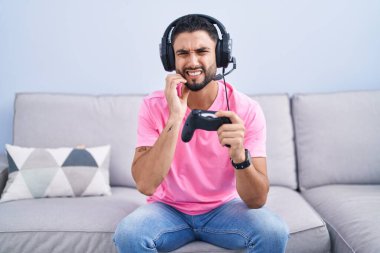 Hispanic young man playing video game holding controller sitting on the sofa touching mouth with hand with painful expression because of toothache or dental illness on teeth. dentist concept. 