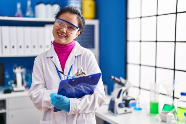 Young chinese woman scientist smiling confident write on clipboard at laboratory