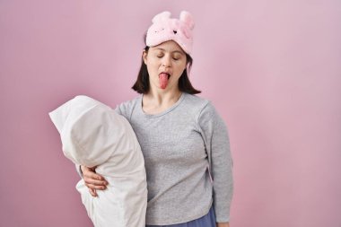 Woman with down syndrome wearing sleeping mask hugging pillow sticking tongue out happy with funny expression. emotion concept. 