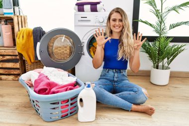 Young beautiful woman doing laundry sitting by wicker basket showing and pointing up with fingers number nine while smiling confident and happy. 