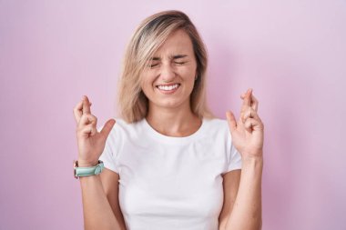 Young blonde woman standing over pink background gesturing finger crossed smiling with hope and eyes closed. luck and superstitious concept. 