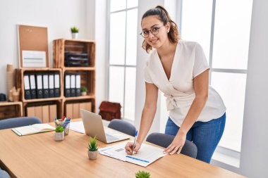 Young beautiful hispanic woman business worker writing on document standing at office
