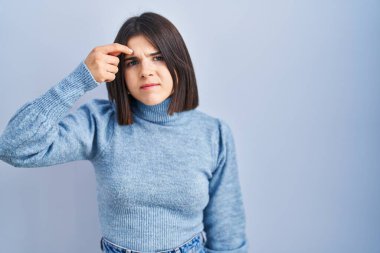 Young hispanic woman standing over blue background pointing unhappy to pimple on forehead, ugly infection of blackhead. acne and skin problem 