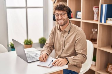 Ofiste çalışan genç bir iş adamı dizüstü bilgisayar ve kulaklık kullanıyor.