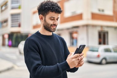 Genç Arap adam sokakta akıllı telefon kullanarak kendinden emin gülümsüyor.