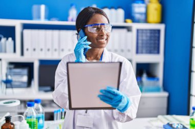 Young african american woman scientist talking on the smartphone reading document at laboratory