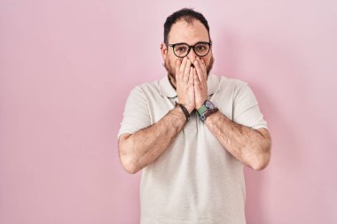 Plus size hispanic man with beard standing over pink background laughing and embarrassed giggle covering mouth with hands, gossip and scandal concept 