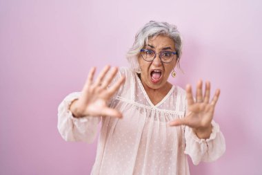 Middle age woman with grey hair standing over pink background afraid and terrified with fear expression stop gesture with hands, shouting in shock. panic concept. 