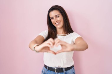 Genç esmer kadın pembe arka planda duruyor. Sevgiyle gülümsüyor. Elleriyle kalp sembolü yapıyor. romantik kavram. 