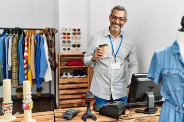 Middle age grey-haired man shop assistant using computer drinking coffee at clothing store
