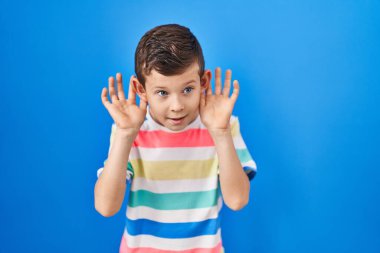 Young caucasian kid standing over blue background trying to hear both hands on ear gesture, curious for gossip. hearing problem, deaf 