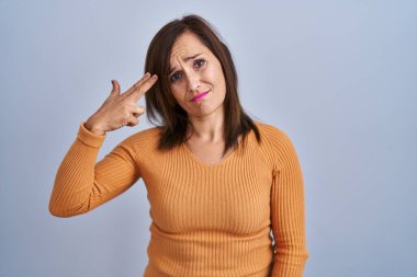 Middle age brunette woman standing wearing orange sweater shooting and killing oneself pointing hand and fingers to head like gun, suicide gesture. 