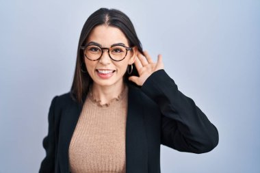 Young brunette woman standing over blue background smiling with hand over ear listening an hearing to rumor or gossip. deafness concept. 