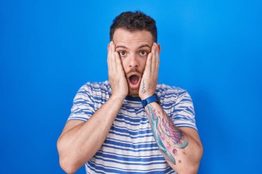 Young hispanic man standing over blue background afraid and shocked, surprise and amazed expression with hands on face 