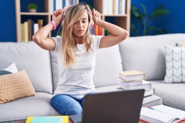 Young blonde woman studying using computer laptop at home doing funny gesture with finger over head as bull horns 
