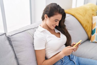 Evdeki kanepede oturan, akıllı telefon kullanan genç İspanyol kadın.