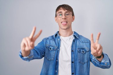 Caucasian blond man standing wearing glasses smiling with tongue out showing fingers of both hands doing victory sign. number two. 