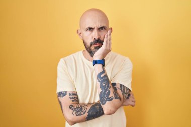 Hispanic man with tattoos standing over yellow background thinking looking tired and bored with depression problems with crossed arms. 