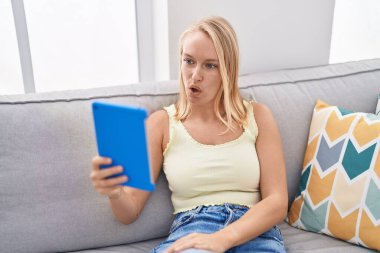 Young caucasian woman using touchpad sitting on the sofa scared and amazed with open mouth for surprise, disbelief face 