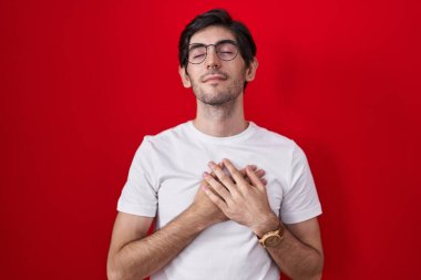 Kırmızı arka planda duran genç İspanyol adam elleri göğsünde, gözleri kapalı ve yüzünde minnet dolu bir jest ile gülümsüyor. sağlık kavramı. 