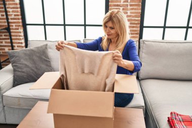 Middle age blonde woman sitting on the sofa opening a box with sweater skeptic and nervous, frowning upset because of problem. negative person. 
