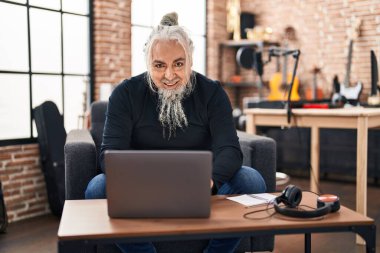 Middle age grey-haired man musician using laptop sitting on chair at music studio