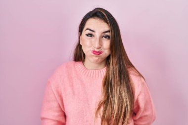Young hispanic woman standing over pink background puffing cheeks with funny face. mouth inflated with air, crazy expression. 