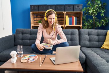 Young beautiful woman practicing painting looking at online tutorial looking positive and happy standing and smiling with a confident smile showing teeth 