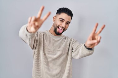 Young handsome man standing over isolated background smiling with tongue out showing fingers of both hands doing victory sign. number two. 