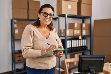 Middle age hispanic woman ecommerce business worker writing on clipboard at office