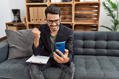 Young hispanic psychologist man doing therapy on video call with smartphone screaming proud, celebrating victory and success very excited with raised arms 