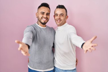 Homosexual couple standing over pink background looking at the camera smiling with open arms for hug. cheerful expression embracing happiness. 